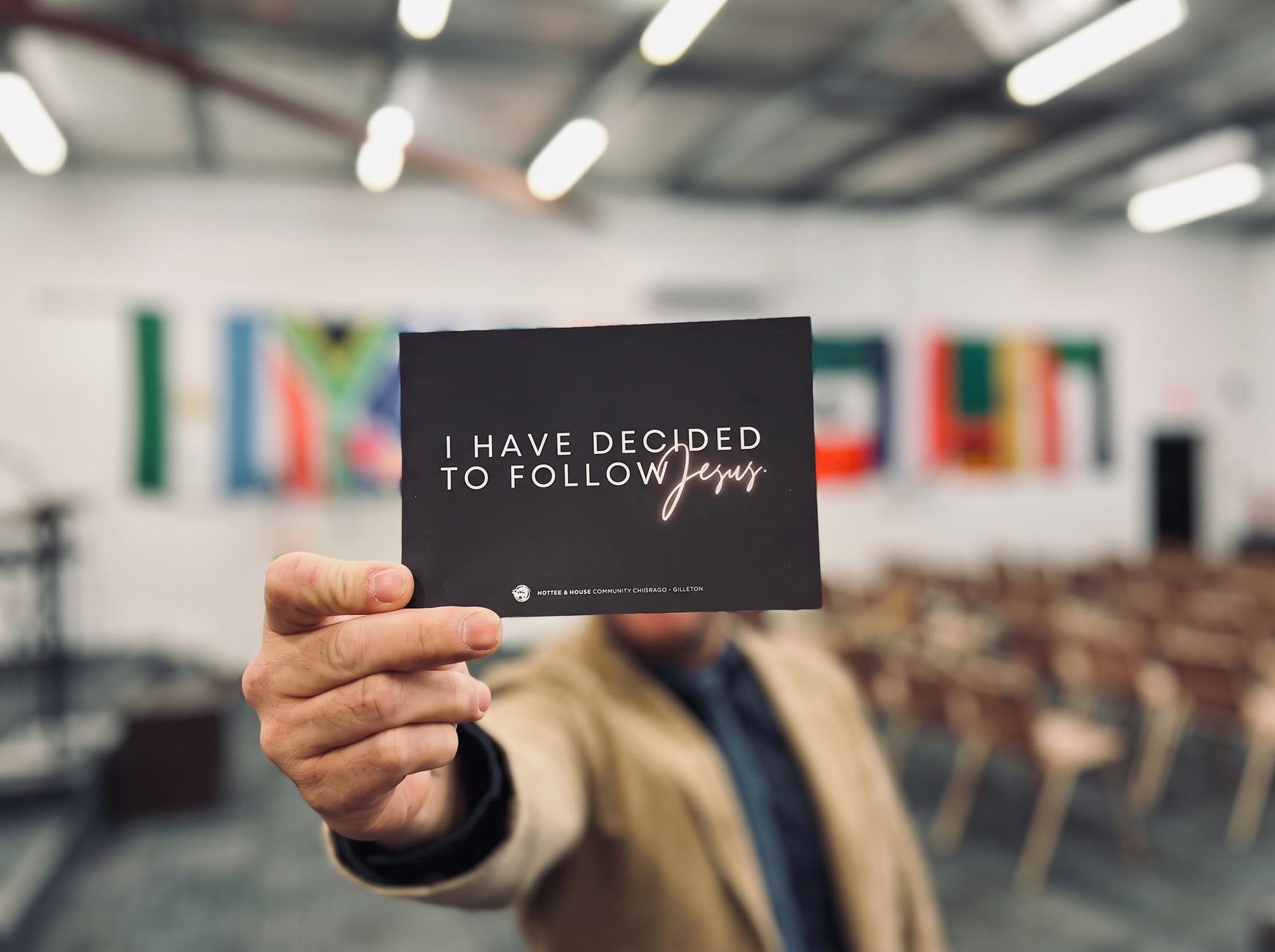 A hand holding a card that reads 'I have decided to follow Jesus' in the church hall, with flags of many nations on the wall